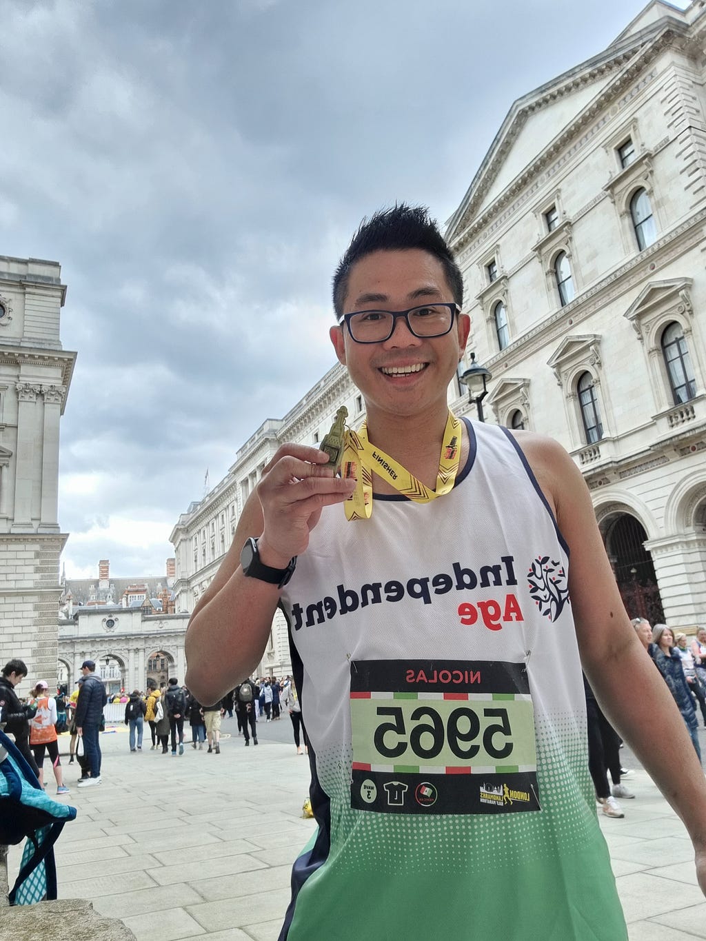 Nicolas, a man with short dark hair and glasses, smiles at the camera while wearing an Independent Age running vest and holding up a yellow running medal he’s been awarded.