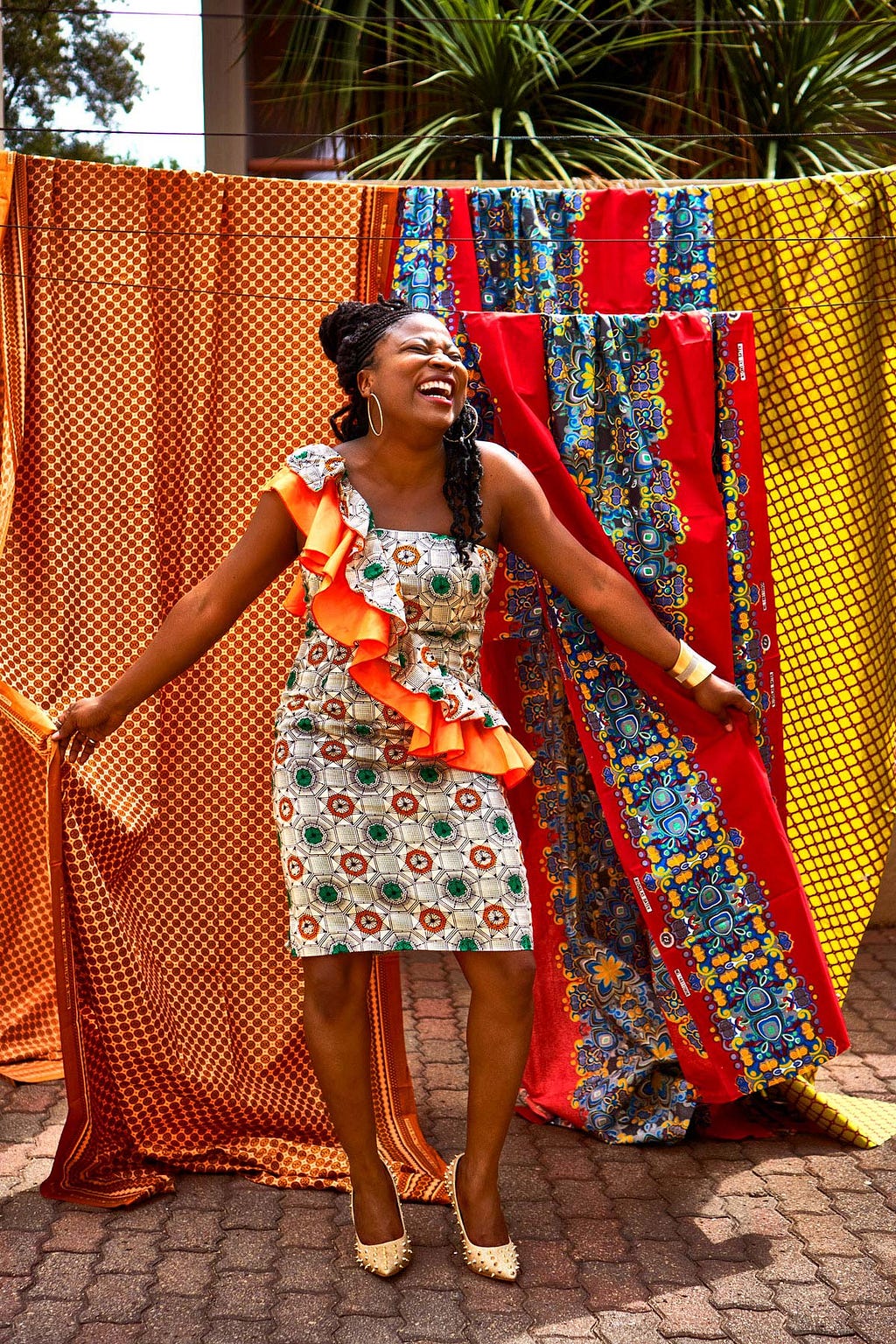 Victoria Adasonla, wearing a colorful traditional kaftan, stands in front of an array of traditional African fabrics.