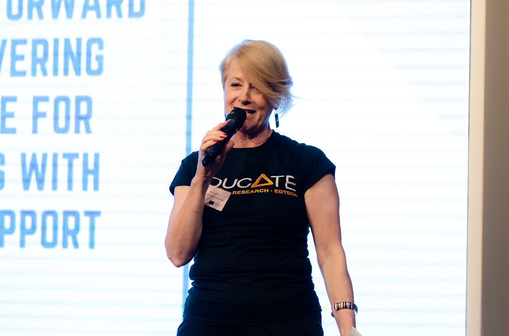 Professor Rose Luckin speaks through a microphone backlit by a huge white light screen.