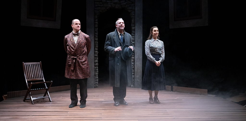 Three actors stand onstage and prepare to take a bow; the actor on the left wears a patterned smoking jacket, the one in the middle a gray suit, and the one on the right a blue skirt and floral blouse.