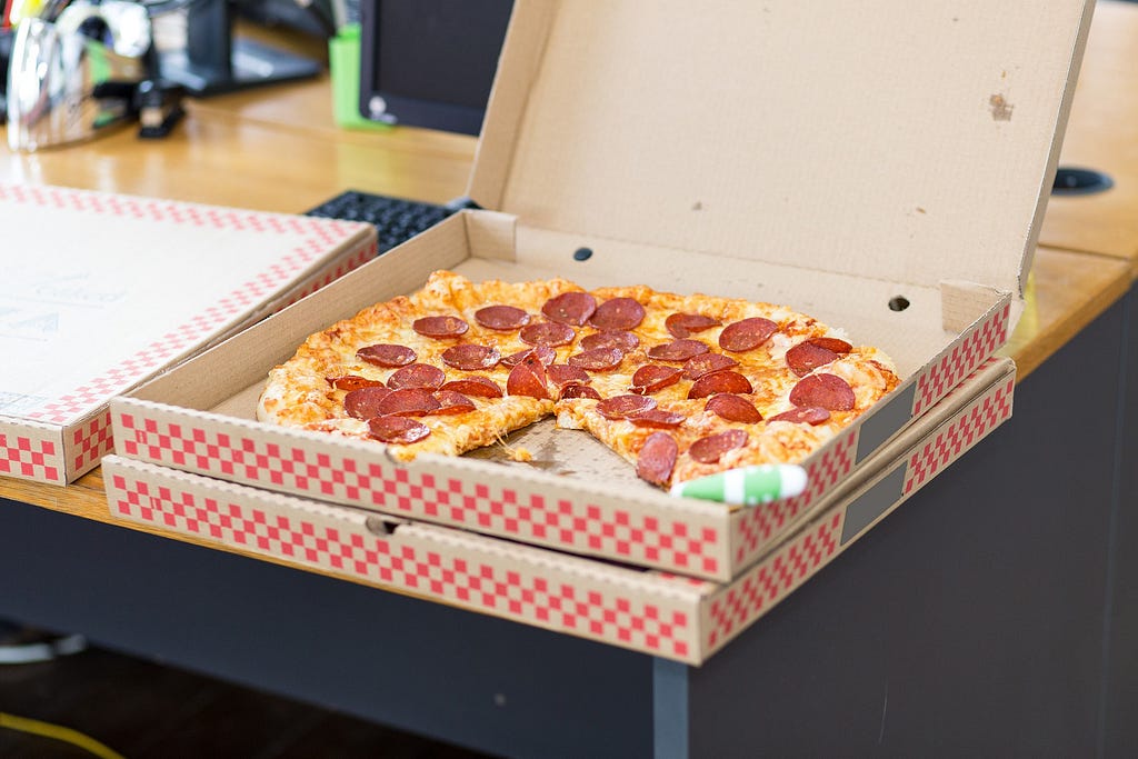 Two pizza boxes are stacked on a table. The top box is open revealing a pepperoni pizza with a slice removed.