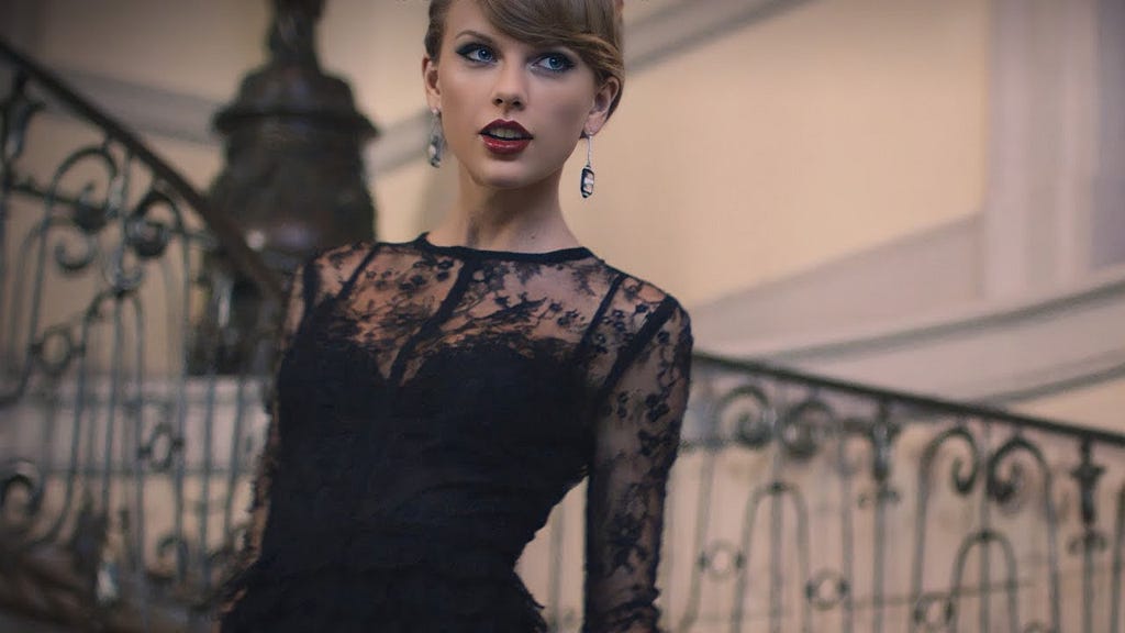 Picture of Taylor Swift in the ‘Blank Space’ music video. She’s a white woman with blonde hair wearing long earrings, red lipstick and a black lace dress and standing on a staircase.