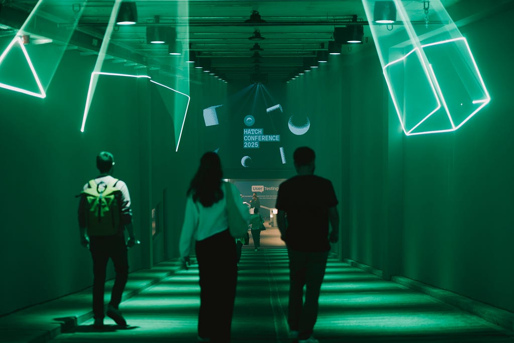 Three people, pictured from behind, walking down a hallway towards the entrance to Hatch Conference 2025, with neon green lights and projections everywhere.