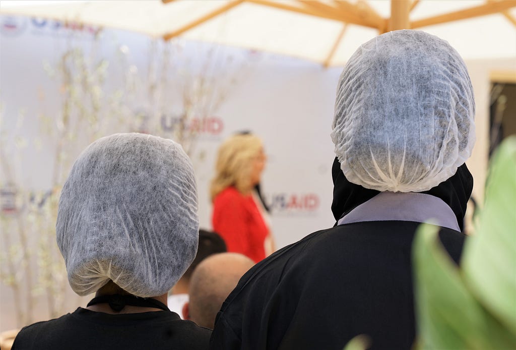 Two Moroccan women dressed in black with hair nets stand as they observe an American woman speaking at a podium.