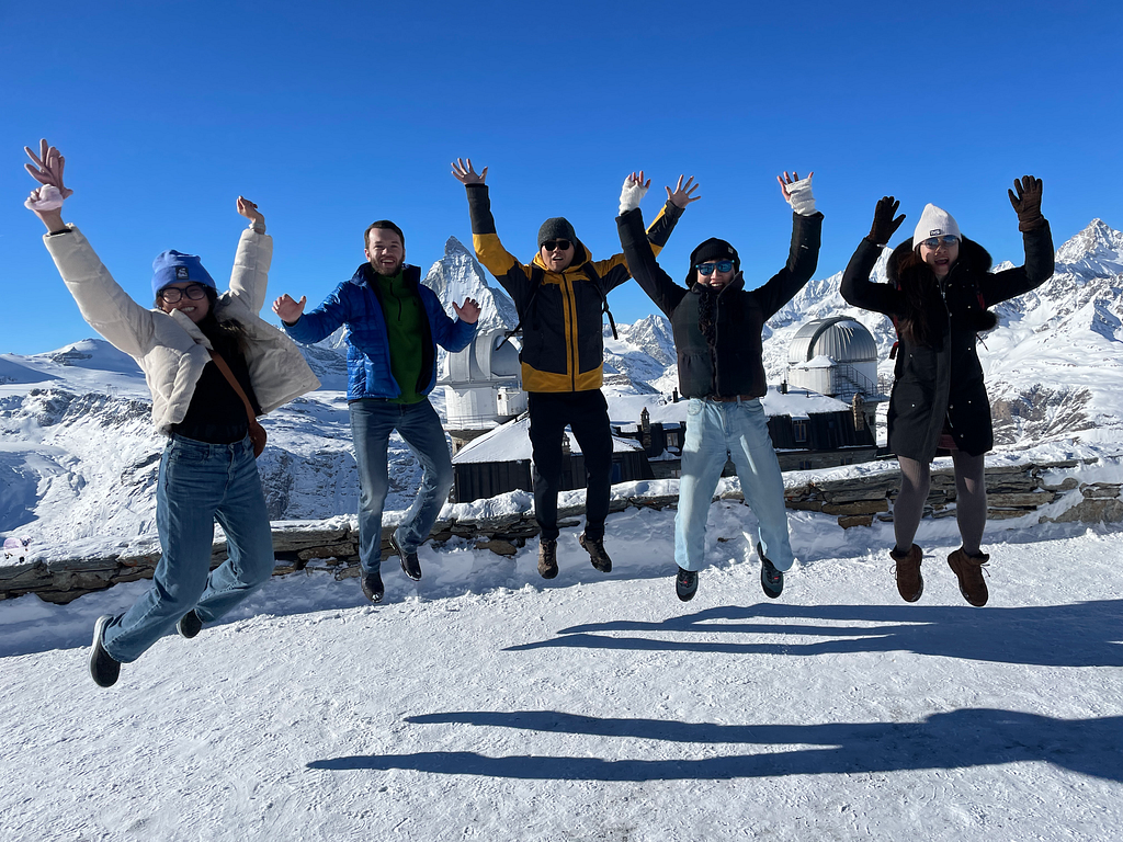 Five people simultaneously jump into the air, with a snowy foreground and white snow-covered mountains and blue skies in the background.