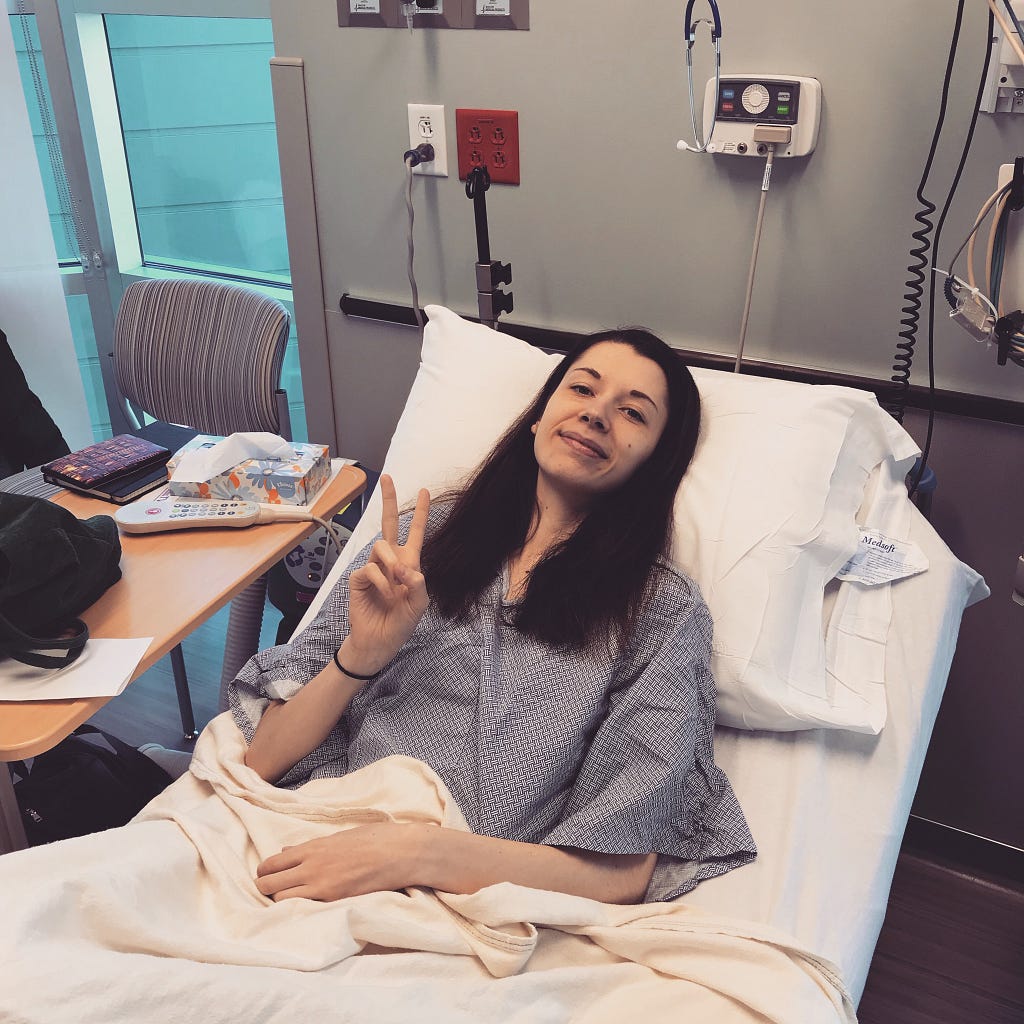 Summer, a woman in her early twenties, smiles from a hospital bed. She wears a gown and her hand makes a “peace” sign.