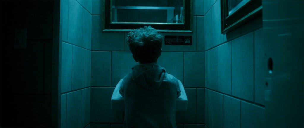 A man stands at a sink in a blue-lit bathroom. He has blonde hair and is wearing a gray hoodie.