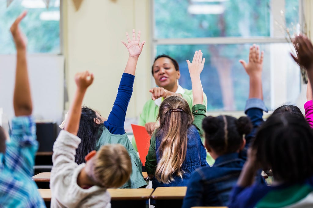 A teacher calls on a student, sitting among several elementary-aged students with their hands raised.