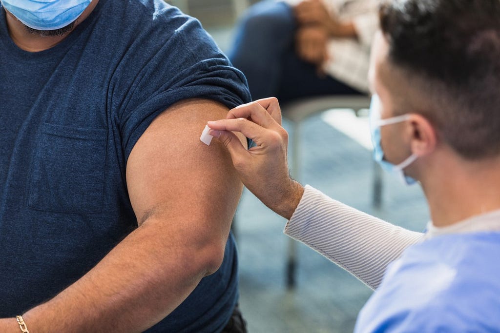 Una imagen de un hombre con una camisa azul marino con la manga remangada, a punto de recibir la vacuna en el brazo izquierdo de un enfermero.