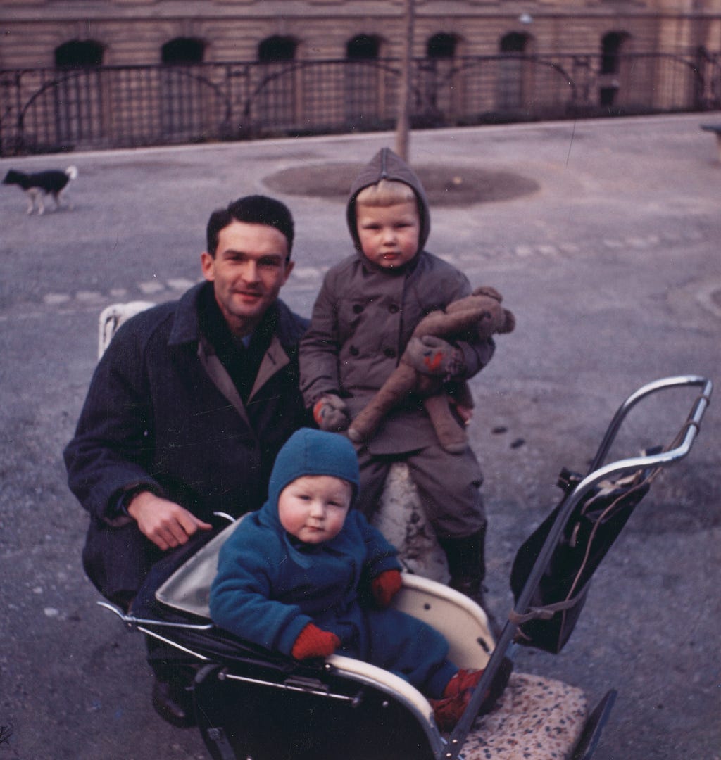 A man crouches beside two young boys with rosy cheeks, one wearing a brown coat and hood and holding a stuffed animal, the other sitting in a stroller and wearing a blue coat and hood. There is a dog in the far left corner of the photo.