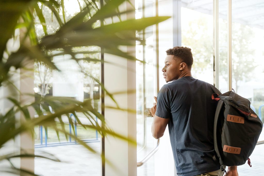 A man in a blue t-shirt with a black backpack exiting an office building.