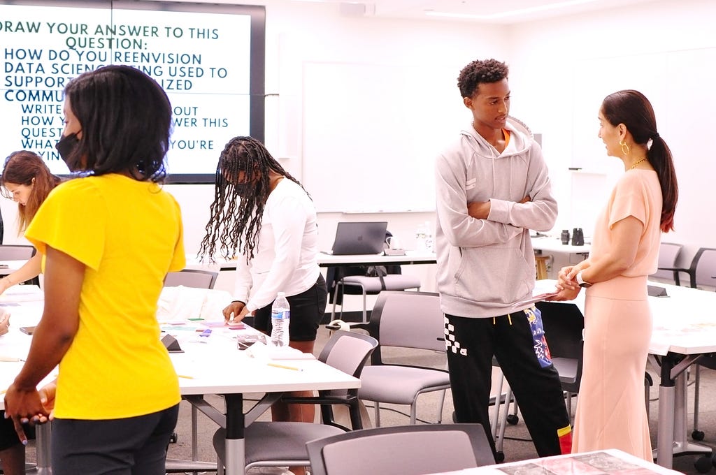 Photo of an MIT classroom with a screen on the wall displaying the prompt “How do you reenvision data science being used to support marginalized communities?” One student works on her project at a table with the instructor Raechel Walker and undergraduate researcher Zeynep Yalcin on either side of her. Another student stands nearby, talking to Cynthia Breazeal.