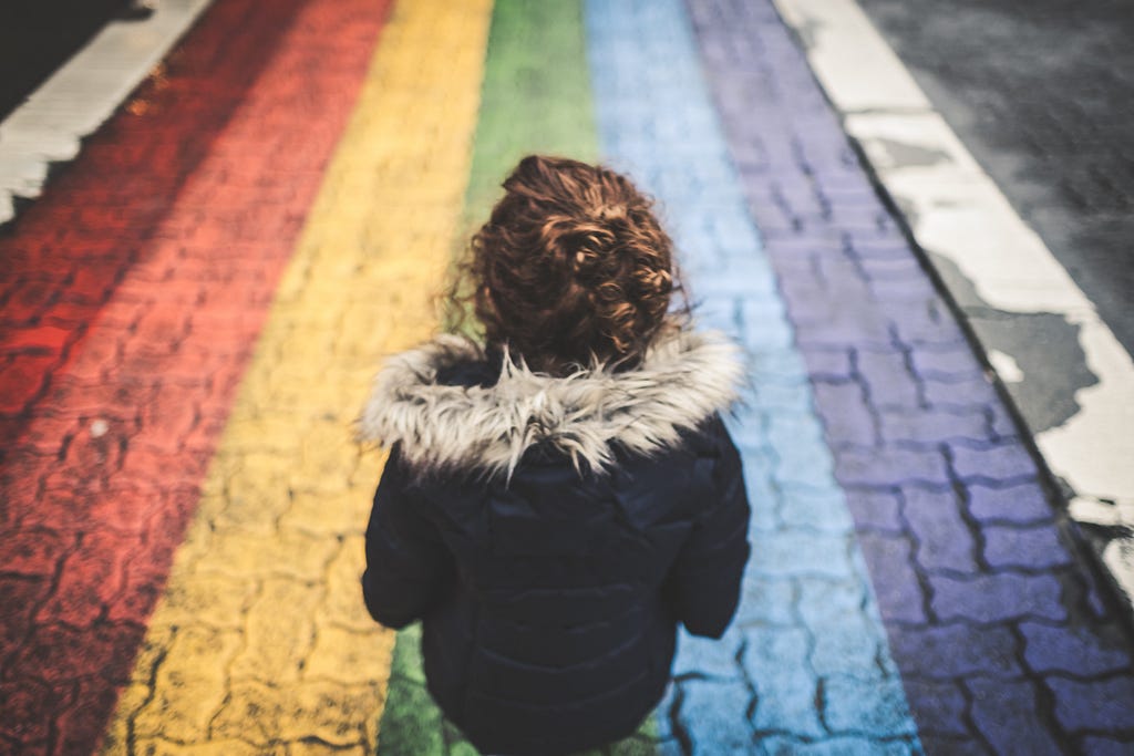 A woman in a warm coat standing on a rainbow coloured road. Photo by Cory Woodward on Unsplash.