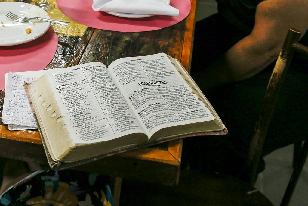 The image shows an open book, specifically a Bible, with the pages opened to the Book of Ecclesiastes. The Bible is placed on a wooden table with some other items visible, including a plate with remnants of food, a napkin, and some handwritten notes. The scene suggests a study or reading session, possibly during a meal.
