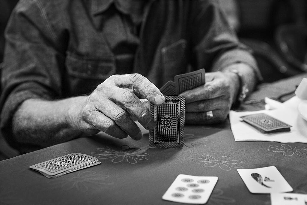 Two wrinkled hands hold a deck of cards. Some cards are face-up on the table. Stefani’s grandfather is playing a game of the Italian card game scopa.