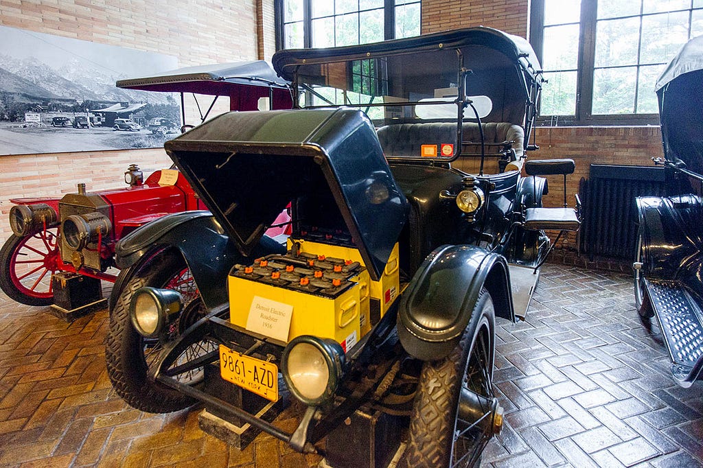 A black classic car with its hood open, revealing a box of batteries. The car is on display in a museum, with a painting of a mountain in the background.