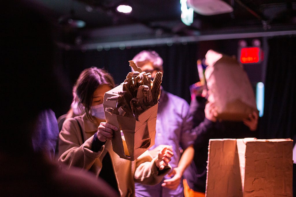 In a dark room, a participant examines a cardboard mask with a triangular nose and textured tufts of hair standing up. In the background, people try other masks.