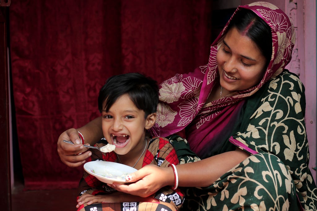 A woman holds a small bowl of yogurt in one hand and a spoon of yogurt in the other as a smiling child opens her mouth wide and leans in for a mouthful.