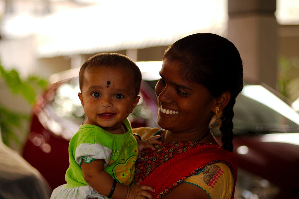 A woman wearing a red and yellow sari holding an infant, smiling.