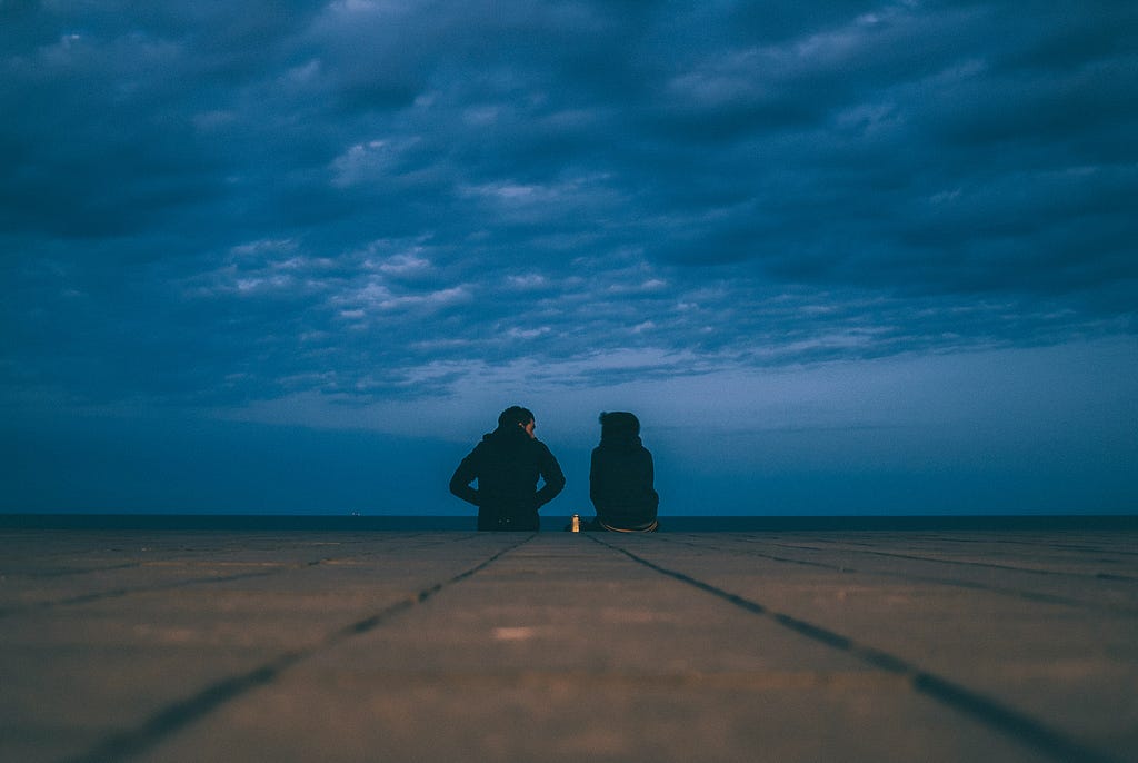 Duas pessoas sentadas conversando em frente ao mar em um lugar frio.