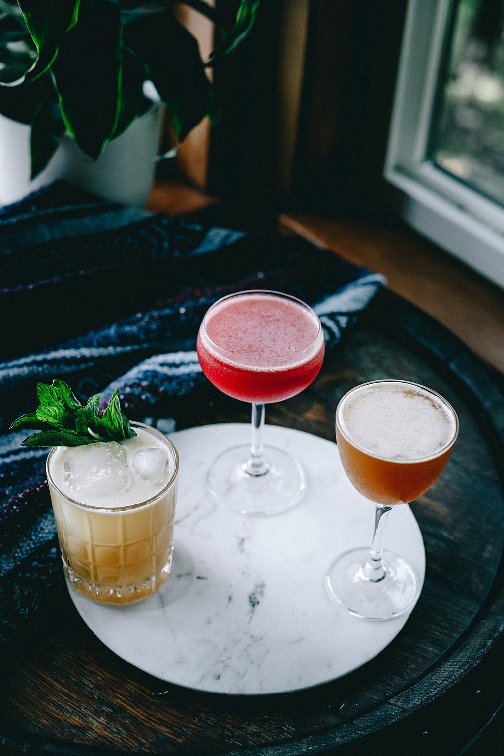 Three cocktails sitting on a white, marble circle. Cocktail on the left is gold in colour with a sprig of mint coming out the top, middle is a deep red cocktail in a coupe glass, and the right is a syrup-coloured cocktail in a nick and nora glass