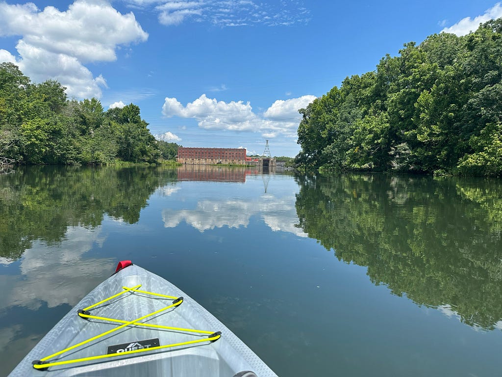 Front of grey kayak heading towards a brick building at the end of dam. Two tree lined shores with smooth reflective water.