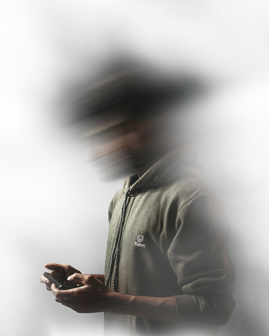 Waist-up image of a medium-dark brown skinned man wearing an olive green hoodie and holding his phone in both hands. His hands and the front of his torso are in full focus while the rest of him is severely blurred into the white and light-grey background — the overall effect is somewhat ghostly.
