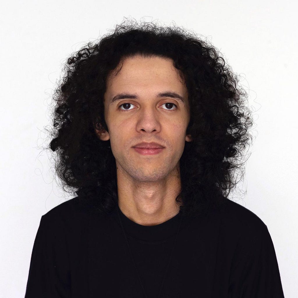 A light-skinned man with long wavy black hair, black eyes, and thick lips, wearing a black t-shirt, against a white background.