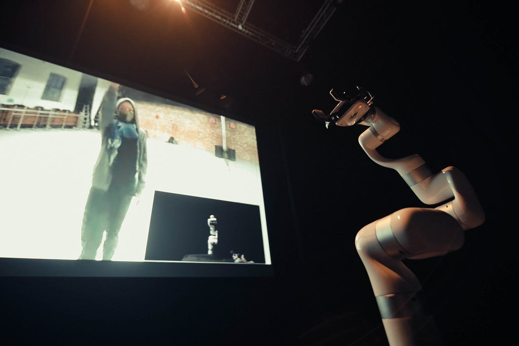 A woman is dancing on the screen on the left while a robot arm looks on from the right.