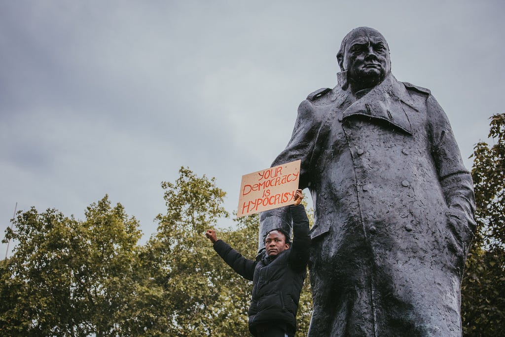 Image of man standing in front of a statue of Churchill holding a sign that reads “Your democracy is hypocrisy”.