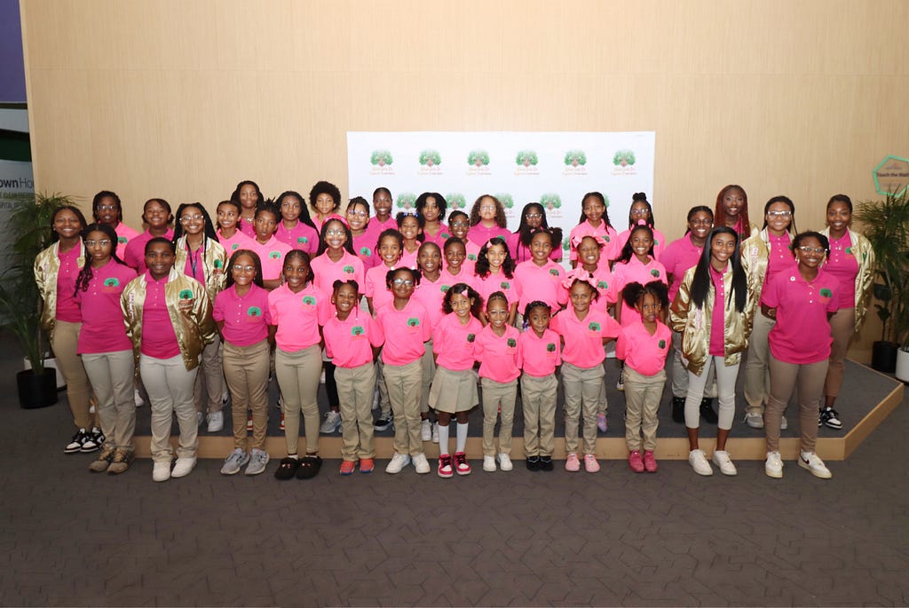 A group of Black Girls Do Engineer members standing together to take a group picture. All of the young girls are wearing pink shirts with a Black Girls Do Engineer logo and khaki pants. The girls have a diverse set of hairstyles including braids, ponytails, straightened hair, afros, and curly hair. The group of girls are standing in front of a tan wall with a white poster that has a green Black Girls Do Engineer logos on it.