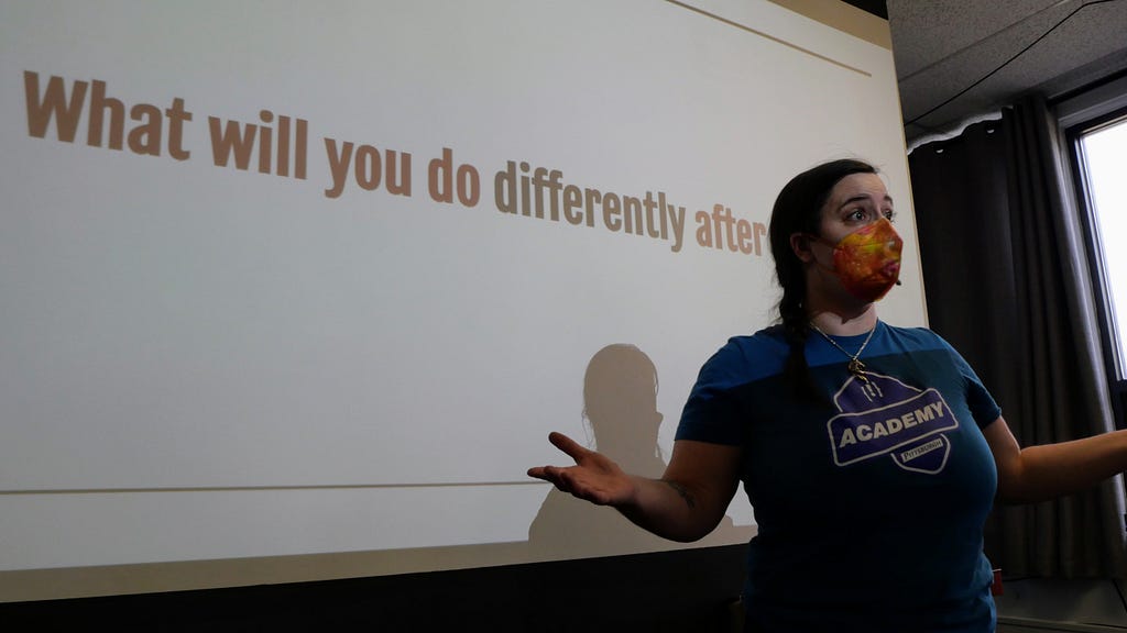 A woman wearing an orange fast mask and a blue T-shirt that says Academy Pittsburgh stands in front of a projected display that says What Will You Do Differently After [Today].