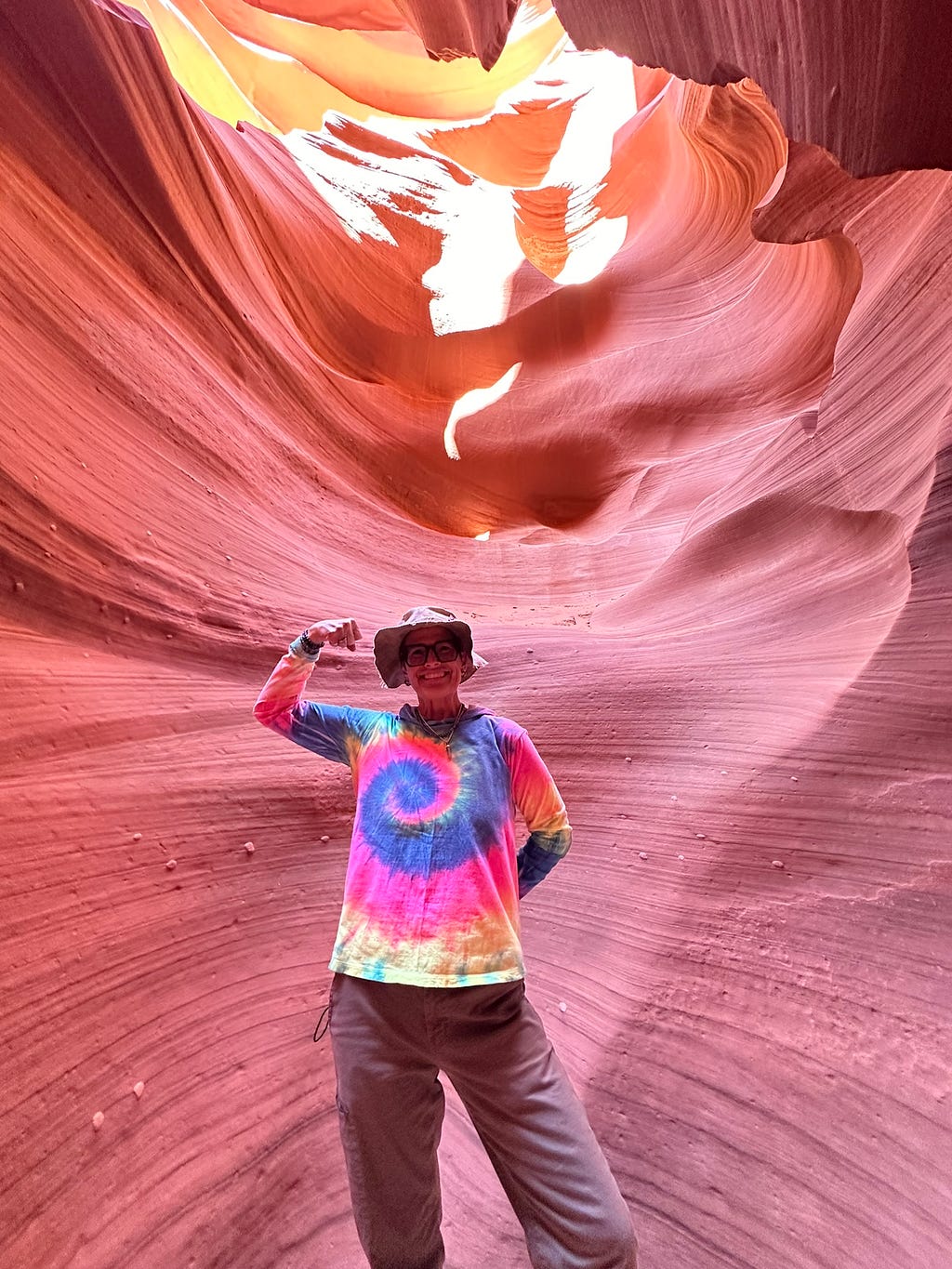 CC appears in a rainbow tie dyed long sleeve hoodie and a tan hiking hat standing in front of one of the orange/red canyon walls of lower antelope canyon and doing the classic colorectal cancer strong arm selfie.