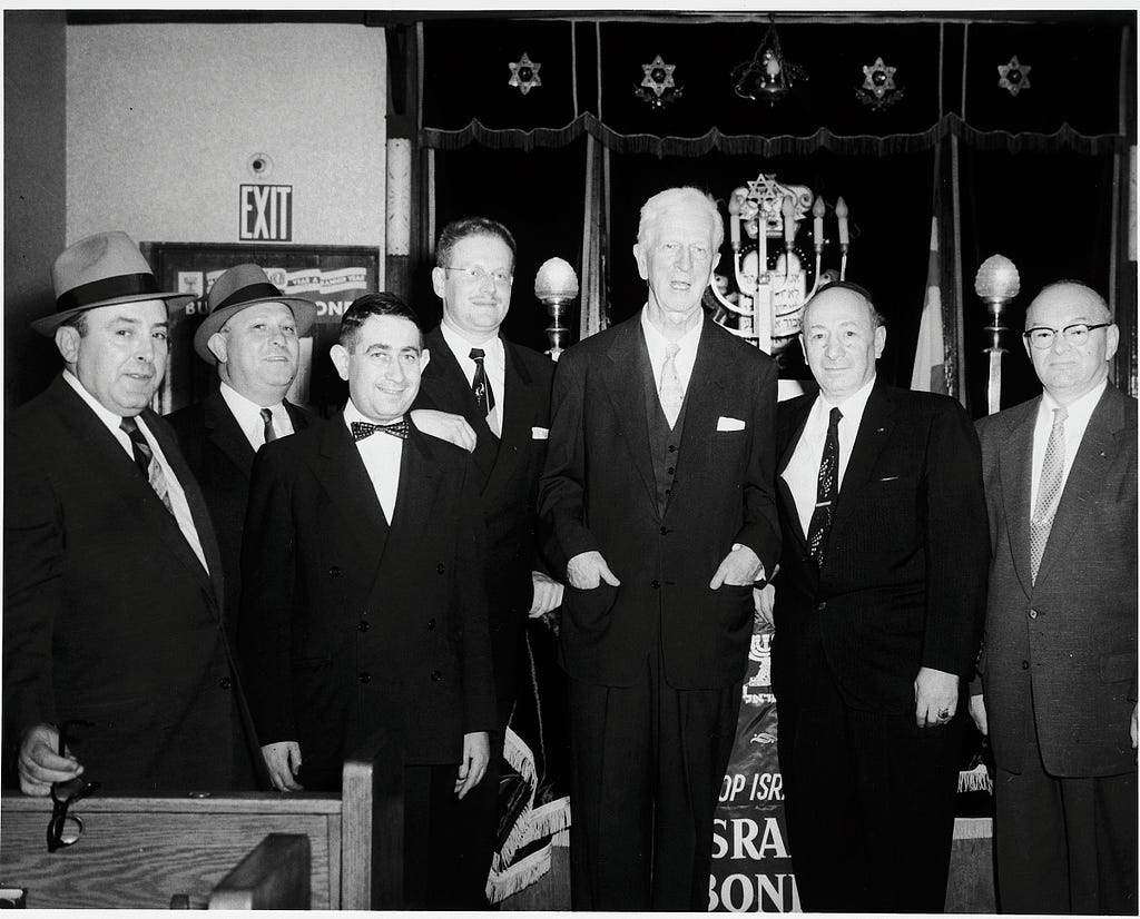 The same man is seen standing with a group of six other men, all in business formal attire. In the background can be seen a menorah, a heavy drape decorated with Stars of David, and an exit sign.