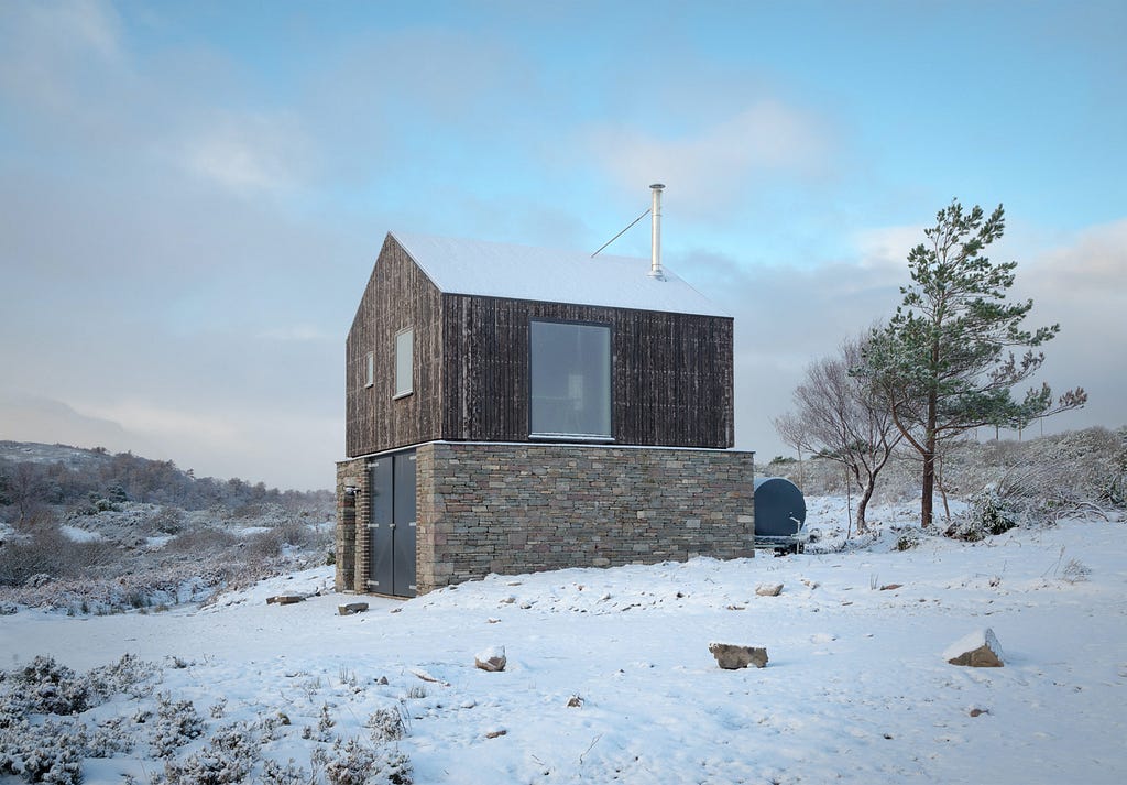 A drystone wall and wooden-clad building sits in a field of snow.