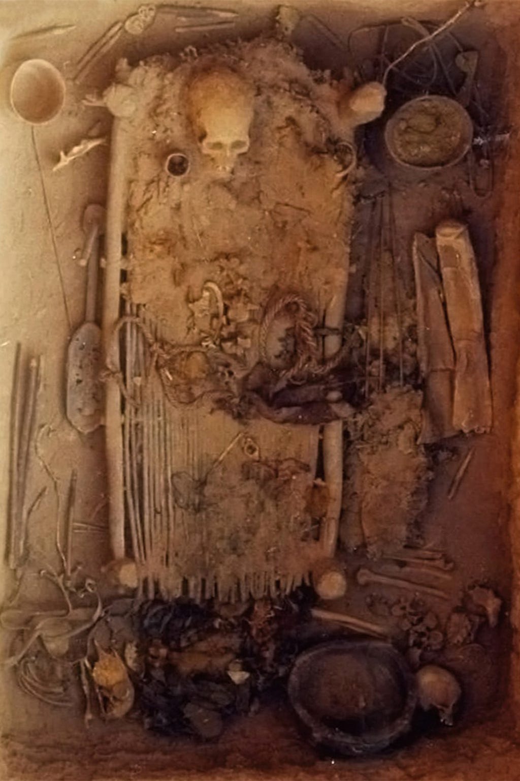 A skeleton rests on a bier made of long sticks in a dry, dusty grave. Various containers and implements surround the bier.