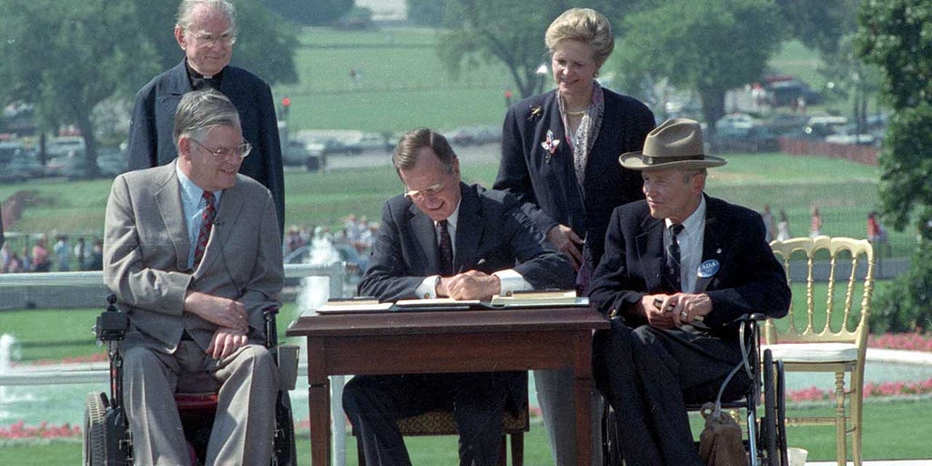 Picture of President George H. W. Bush signing the Americans with Disabilities Act in 1990. The picure is take outside and the President is sitting at a desk. There are four other people in the picture, two of whom are standing behind and on either side of the President (a man and a woman) and two of whom (men) are sitting in wheelchairs on both sides of the desk.