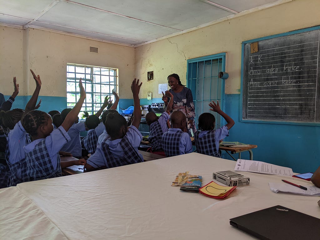 Students raise their hands high into the air as the student teacher at the head of the classroom looks on joyfully.