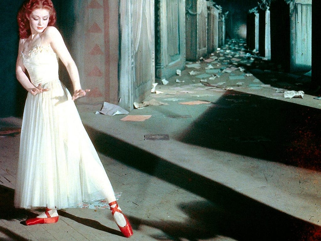 A redheaded woman in a white dress and red shoes standing at the end of a grey corridor.