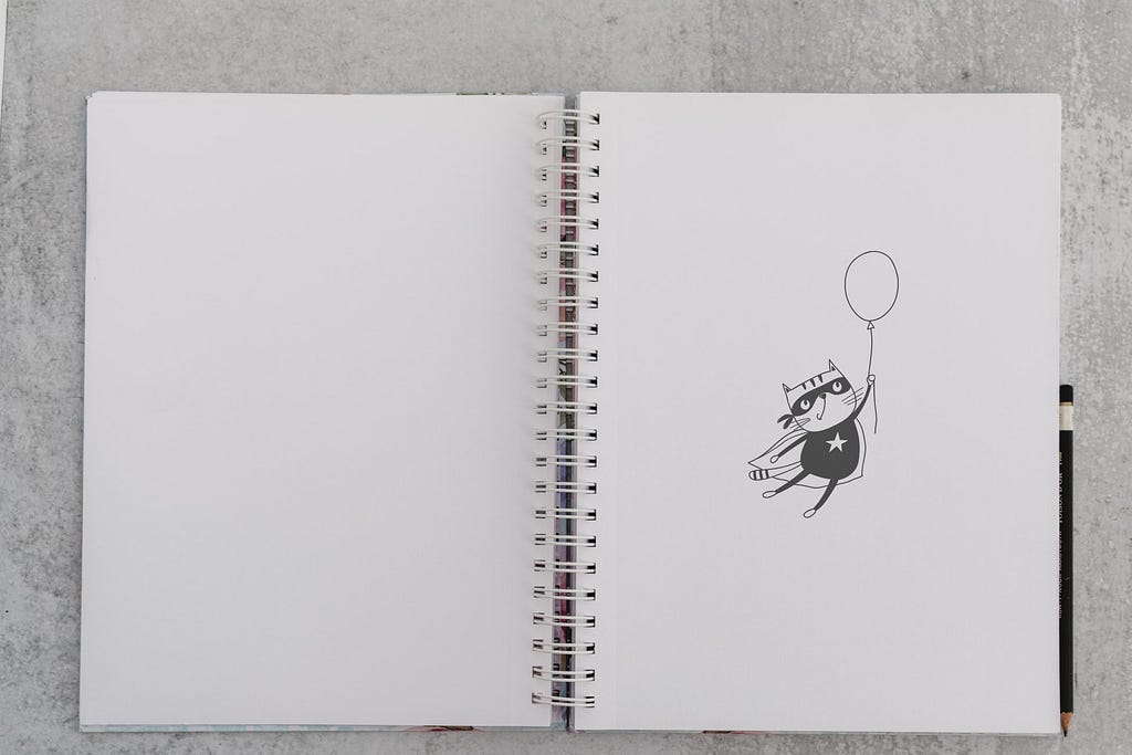 foto de um caderno de folhas brancas com o desenho de um gato vestido de super herói segurando um balão e voando