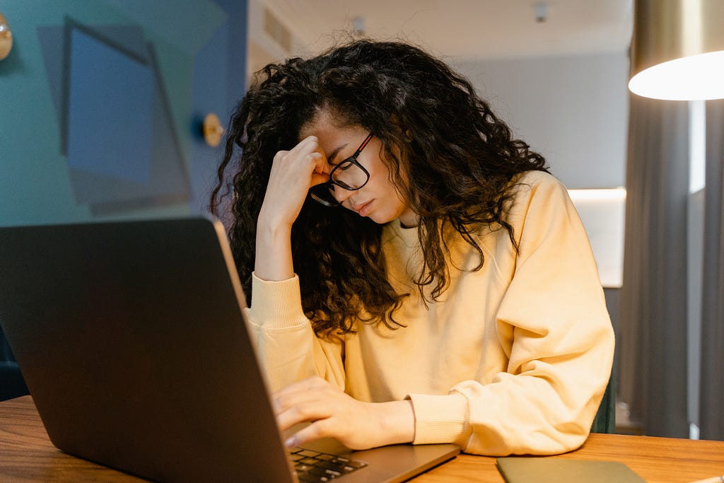 Young woman with dark curly hair holding her head in one hand while typing on her laptop keyboard with the other