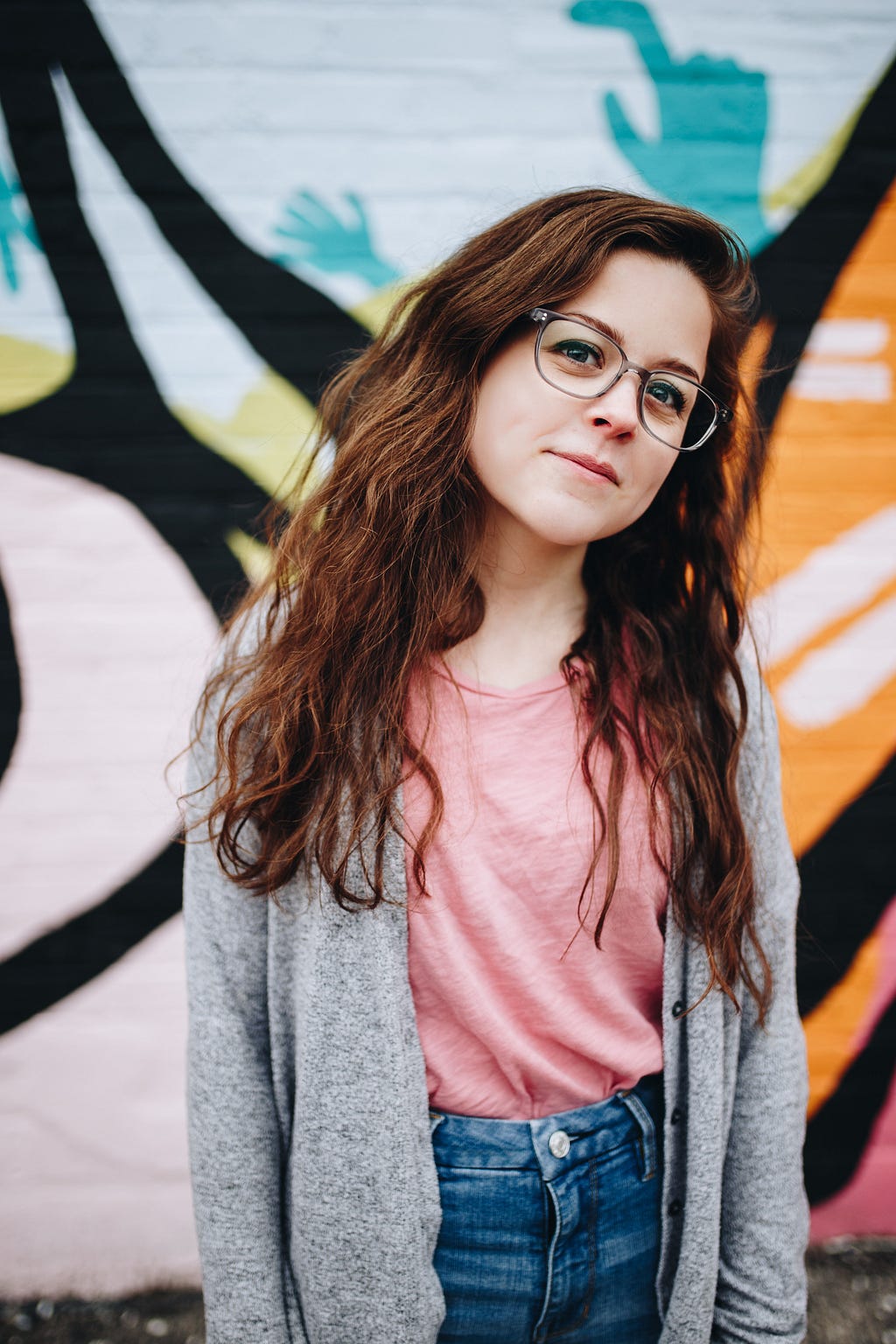 My persona, Ria, long wavy but not quite curly red hair, glasses, dressed in a casual pink t-shirt, jeans and a grey cardigan, standing in front of a very vibrant painted wall.