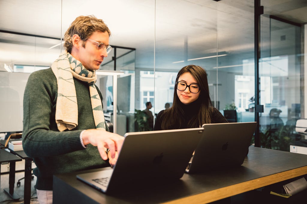 Man and woman stand in front of two laptops on a high table and discuss what is on the screen