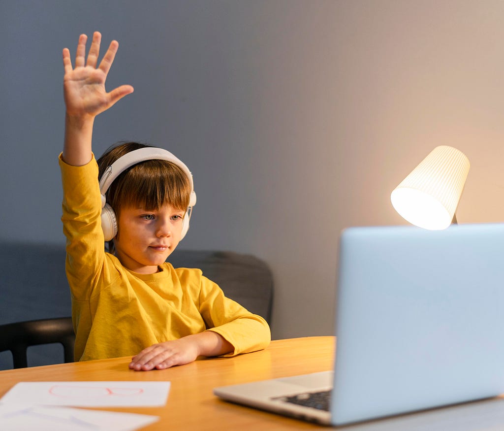 Young boy in a yellow sweater wearing headphones sitting in front of a laptop at a desk raises his hand during his homeschooling lesson