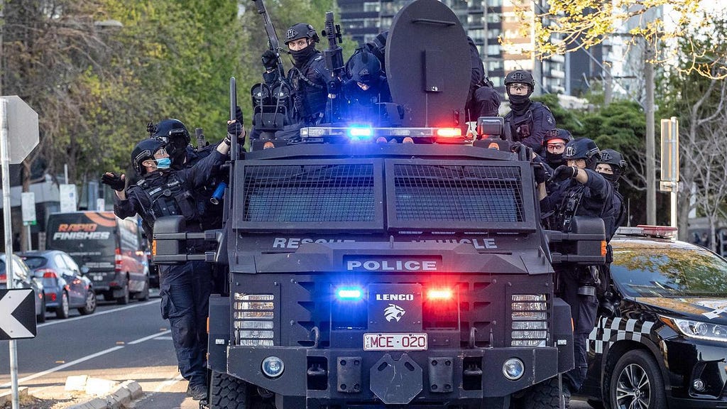 A bulky armoured vehicle labelled ‘police’ with at least ten masked cops clinging to the roof and sides, lights flashing blue and red, an array of exterior technological attachments visible on its roof