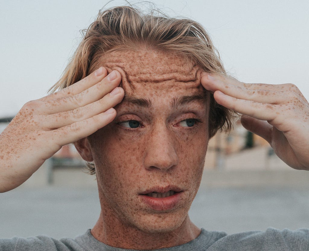 Photo of a man with blond hair and freckles holding the sides of his head looking stressed out.