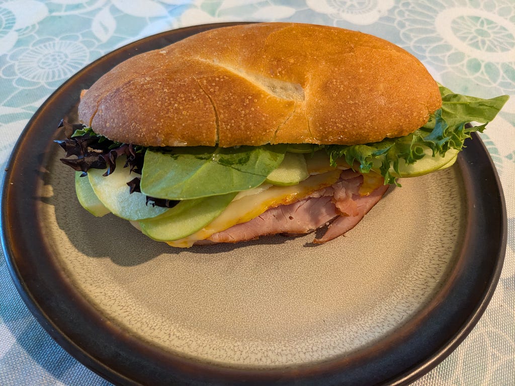 Homemade sandwich containing mixed salad greens, sliced apple, munster cheese and ham on a long sandwich roll, sitting on a beige plate with a brown rim. (There’s mayonaise too but you can’t see it). Tablecloth in background has a blue and white floral pattern.