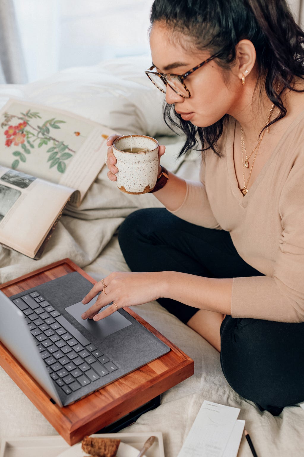 Photo of a woman wearing glasses and a tan sweater sitting cross-legged on a bed. She’s holding a mug of coffee in one hand and using the other hand to navigate a laptop computer.