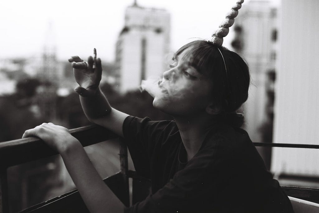 A young lady with dark hair sits on a balcony smoking a cigarette and wearing a unicorn horn. She suits leaning forward with one hand on the rail and one holds her cigarette slightly raised, her elbow rests on the rail. The image is black and white and the background high rise apartments are out of focus.