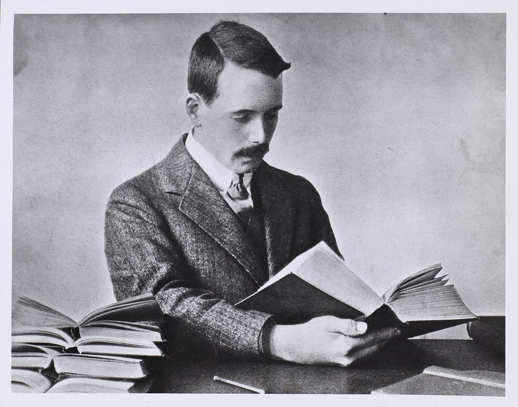 Black and white photo of Henry Gwyn Jeffreys Moseley (1887–1915) sat at a desk reading with a stack of open books piled on the desk beside him